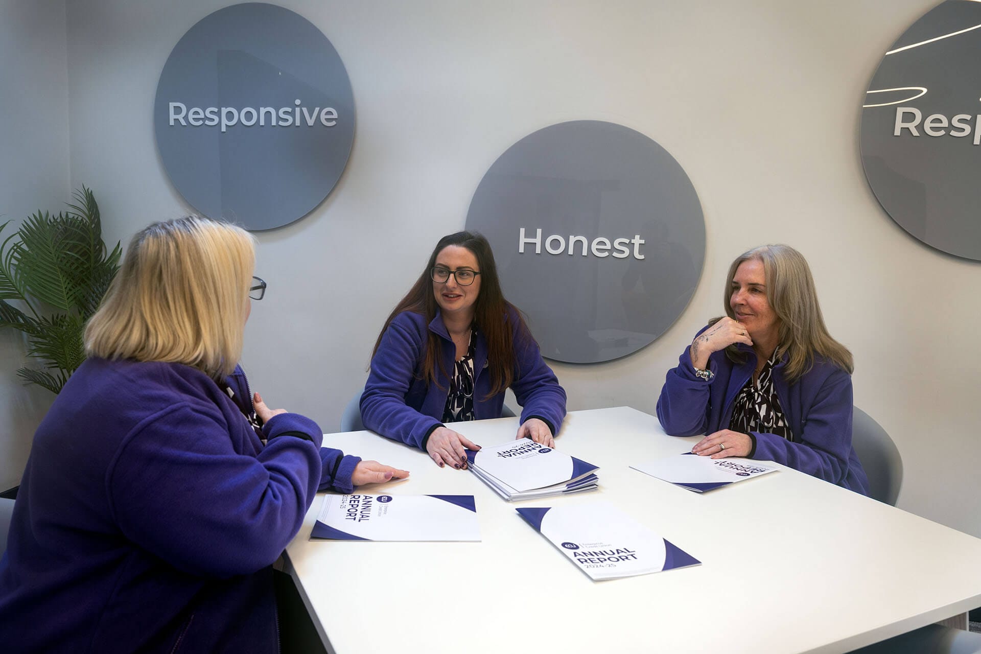 Three women in purple jackets seated around a table in a meeting room with “Responsive” and “Honest” displayed on the wall behind them.