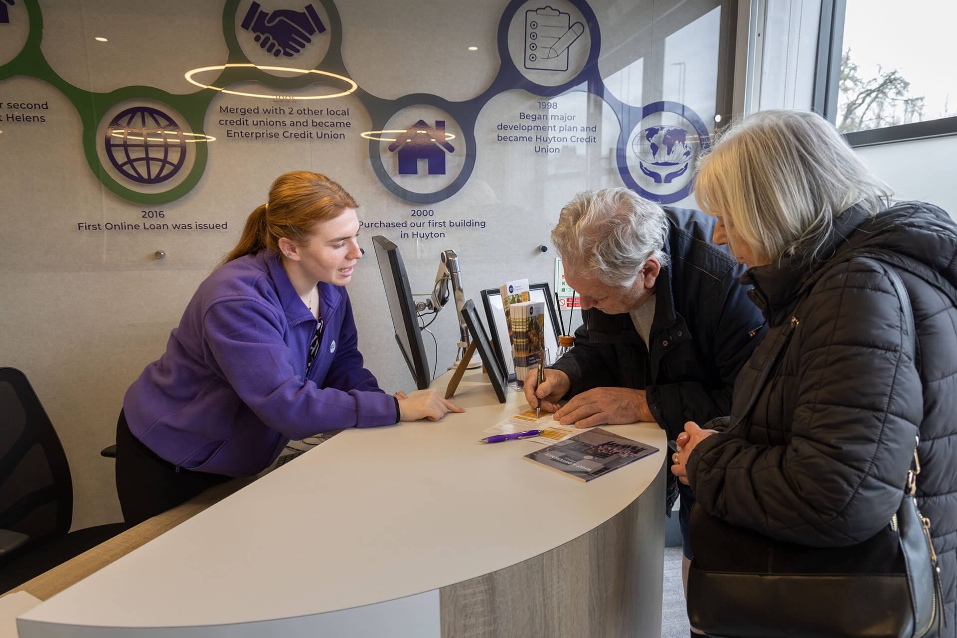 A staff member behind a curved reception desk helping two older visitors fill in paperwork at Enterprise Credit Union.