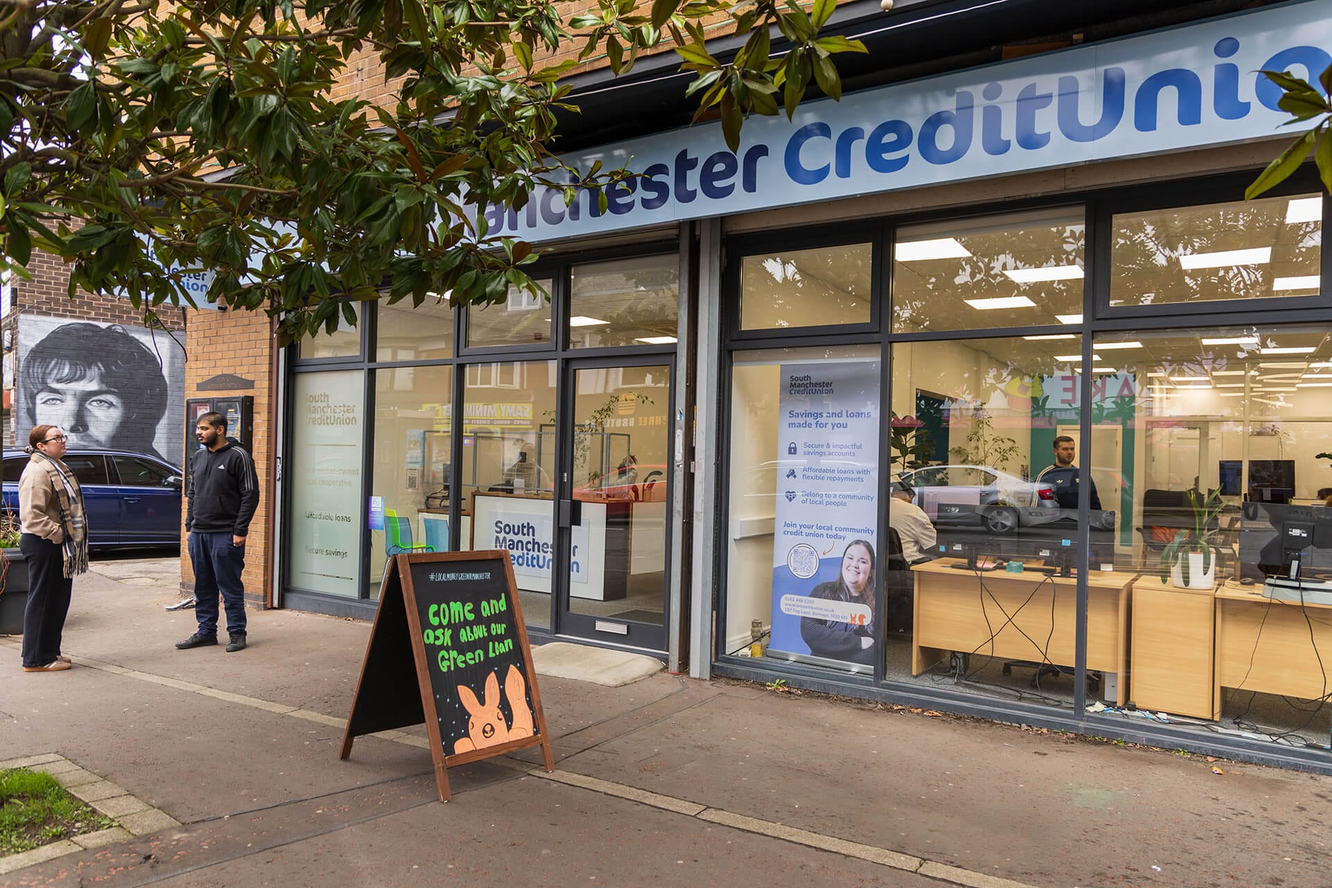 South Manchester Credit Union branch exterior, with two people talking on the pavement outside and staff visible inside through the front windows.